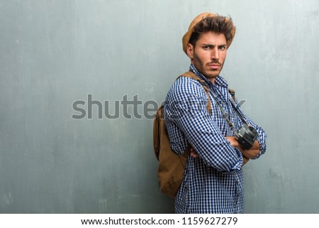 Young handsome traveler man wearing a straw hat, a backpack and a photo camera very angry and upset, very tense, screaming furious, negative and crazy