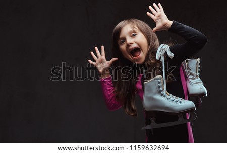 Playful little girl with an amused face dressed in sportswear holds ice skates. Isolated on a dark textured background.