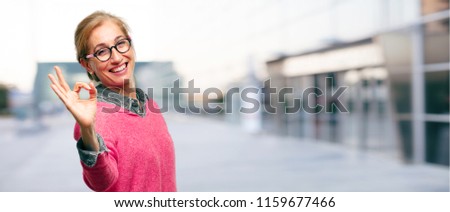 Beautiful adult blonde woman making an "alright" or "okay" gesture approvingly with hand, looking happy and satisfied, with a broad smile. Positive check sign. Lateral or side view.