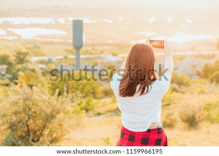 Beautiful girl taking pictures of coutryside from a hill at sunset