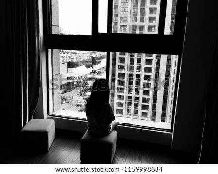 Black and white picture of backside of a kid sitting alone and looking outside through the window. Homesick or sadness or broken family concept