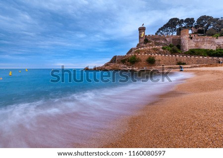 View on small shingle beach with blurred sea wave in front and rocky cape with ancient fortress on top in distance under cloudy sky at evening in Costa Brava (Spain)