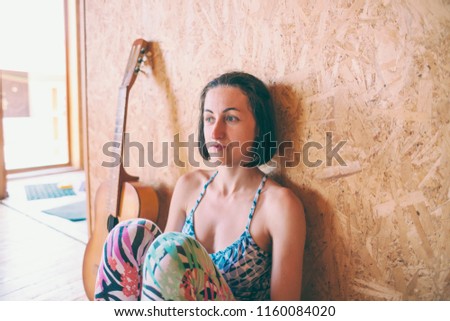 Thoughtful girl sits on the wooden floor next to the guitar. Acoustic guitar. Musician and musical instrument. Woman on the background of a wooden wall.