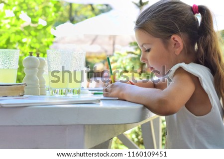 Girl with pony tail hair, drawing outside at the white table with glasses of juice on background