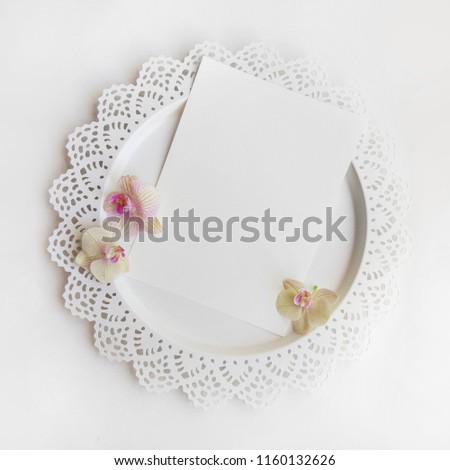 Flat lay composition with orchid flowers, elegant plate and blank paper sheet for text or artwork, white background. Light top view photo for business