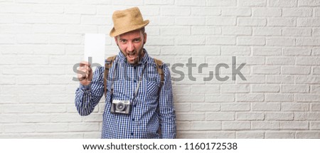 Young traveler man wearing backpack and a vintage camera very angry and upset, very tense, screaming furious, negative and crazy. Holding a white placard.