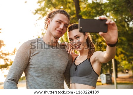 Image of healthy couple man and woman 20s in tracksuits walking in green park together and taking selfie during sunny summer day
