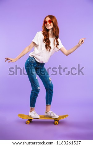 Full length portrait of a smiling young girl standing isolated over violet background, riding on a skateboard