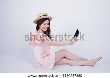 Portrait of a fashionable woman in pink dress and hat sitting on the floor and taking selfie in the studio.
