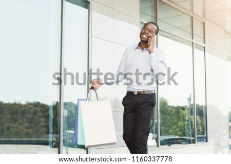 Happy black man with shopping bags talking on mobile, walking along modern boutiques street. Successful business, technology and shopping concept