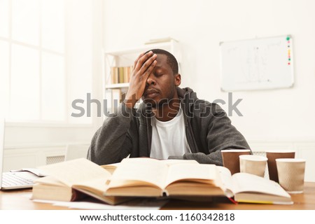 Tired african-american student studying at working table. Exhausted male student preparing for exams, drinking lots of coffee and falling asleep. Education and overworking concept