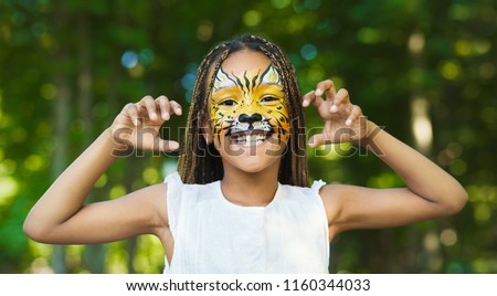 Little african-american girl with tiger face painting roaring, making funny grimace outdoors, copy space Royalty-Free Stock Photo #1160344033