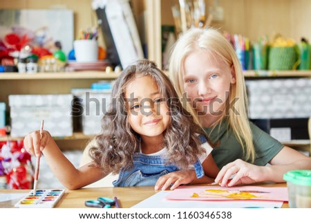 Two sweet girls smiling and looking at camera while painting nice picture during lesson in art school