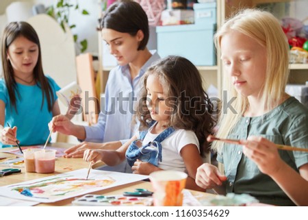 Young female and sweet girls sitting at table in art school and painting bright pictures together