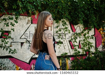 Stylish casual hipster girl in cap, glasses and jeans wear, listening music from headphones of mobile phone against large graffiti wall.