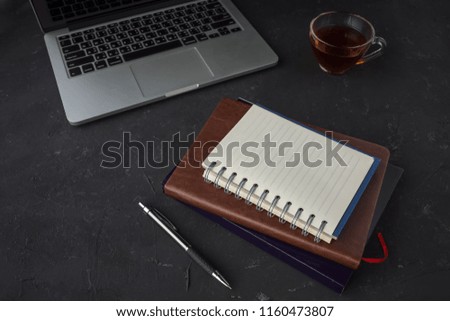 Closeup view of notebook with empty page and a cup of hot tea with laptop on black table. Office desk with accessories. Business or working concept.