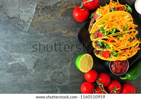 Hard shelled tacos with ground beef, lettuce, tomatoes and cheese. Top view, corner border on a dark background with copy space.