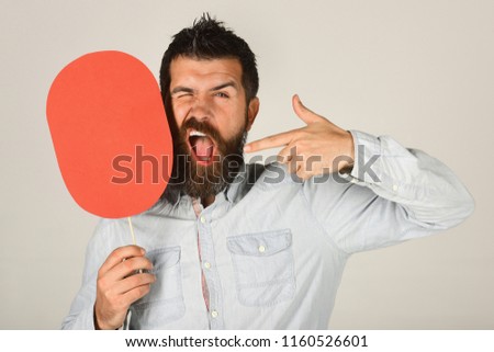 Winking man holds paper nameplate. Bearded man smiling face expression shows finger on red name plate. Man with beard and mustache with funny face winking. Fashion, beauty, hairdressing, barber salon.