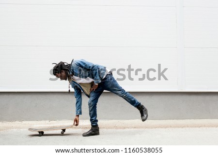 African man with dreadlocks in jeans jacket rides a skateboard on the background of white walls