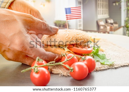 The homemade hamburger with fresh vegetables, french fries, beef, onion, tomato, lettuce, cheese and spices  with little American flags in farmerman hands.