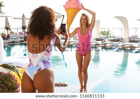 Two pretty young girlfriends taking pictures while posing at the swimming pool resort spa