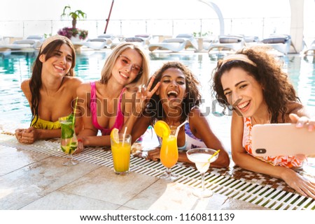 Four happy young women in swimwear taking a selfie while drinking cocktails together at the swimming pool outdoors