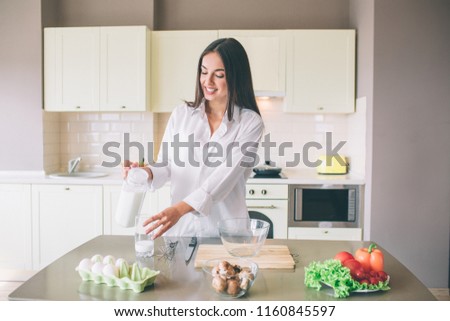 A picture of beutiful girl standing in kitchen and pouring milk in glass cup. She is looking at it and smiling. Girl is concentrated.