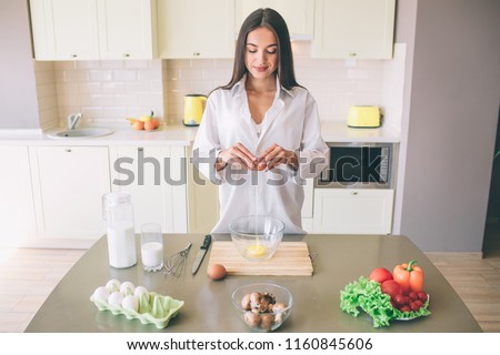 A picture of brunette stands at table with food and looks down. She is pouring egg into big glass bowl. Girl is smiling. She is cooking.