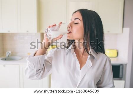 A picture of gorgeus girl standing in kitchen and drinking milk from glass cup. She is keeping eyes closed. Woman wears white shirt.