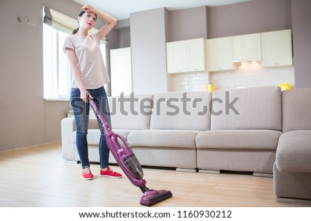 A picture of tired woman stands and holds vacuum cleaner. It is wireless. Girl holds left hand on forehead. She is cleaning the floor.