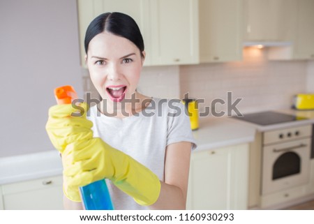 A picture of excited woman stands and holds blue spray in hands. She aims it on camera. Girl stands in kitchen.