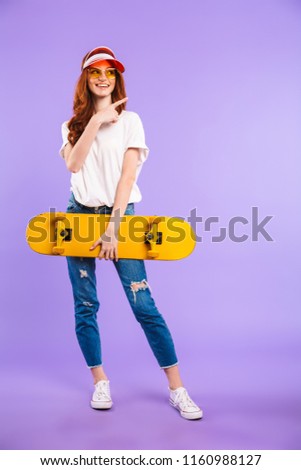 Full length portrait of a cheerful young girl in hat and sunglasses standing isolated over violet background, holding skateboard, pointing away