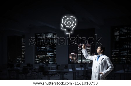 Conceptual image of confident doctor in white uniform interracting with glowing gear brain symbol while standing against night cityscape view on background.