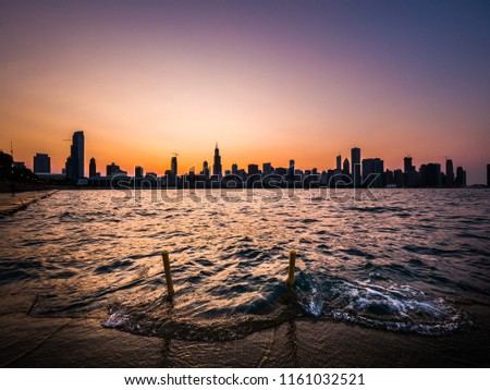 Chicago skyline picture during a beautiful sunset with purple and orange sky above the building silhouettes on the horizon with waves coming up over the concrete shoreline and emergency safety ladder.