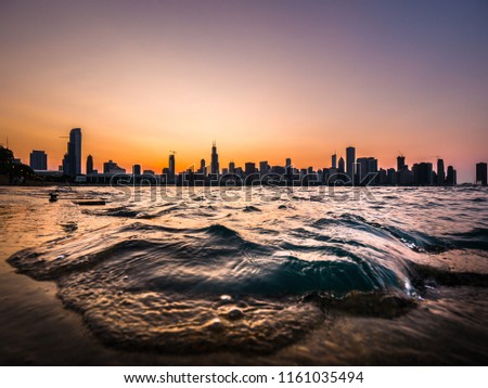 Chicago skyline picture during a beautiful sunset with purple and orange sky above and building silhouettes on the horizon with a wave from Lake Michigan coming up over the concrete shoreline edge.