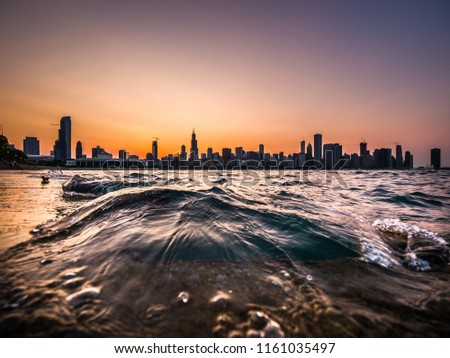 Chicago skyline picture during a beautiful sunset with purple and orange sky above and building silhouettes on the horizon with a wave from Lake Michigan coming up over the concrete shoreline edge.