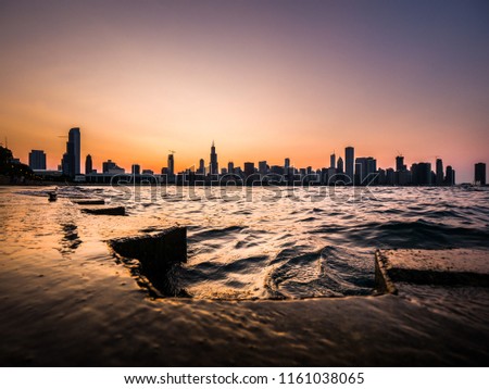Chicago skyline picture during a beautiful sunset with purple and orange sky above and building silhouettes with rippling waves and water of Lake Michigan and concrete shoreline in the foreground.