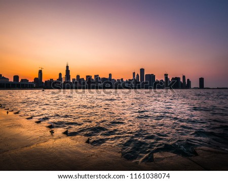 Chicago skyline picture during a beautiful sunset with purple and orange sky above and building silhouettes with rippling waves and water of Lake Michigan and concrete shoreline in the foreground.