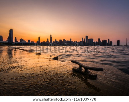 Chicago skyline picture during a beautiful sunset with purple and orange sky above and building silhouettes on the horizon with rippling waves of Lake Michigan and metal boat cleat in the foreground.