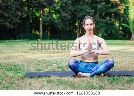 ‎Young woman practicing yoga in nature. Sitting on green grass and meditating, holding hands together. Green background. Healthy and sport concept.