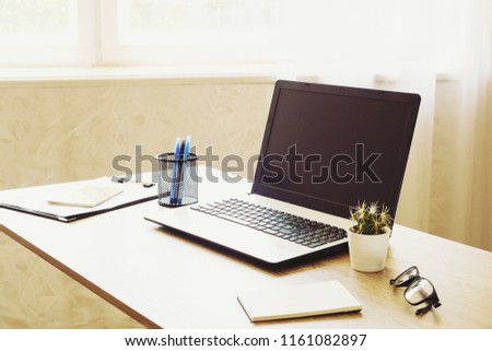 Laptop computer, clipboard, cactus home plant, supplies and folded glasses on wooden desk in spacious office full of sunlight. Designer's creative workspace concept. Close up, copy space, background.