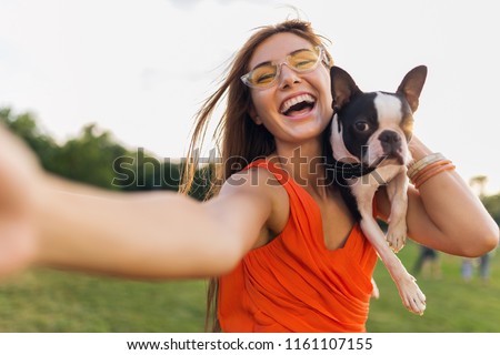 happy pretty woman park making selfie photo on phone camera, holding boston terrier dog, smiling positive mood, trendy summer style, wearing orange dress, sunglasses, playing with pet, having fun