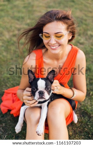 portrait of happy pretty woman sitting on grass in summer park, holding boston terrier dog, smiling positive mood, wearing orange dress, trendy style, sunglasses, playing with pet, having fun