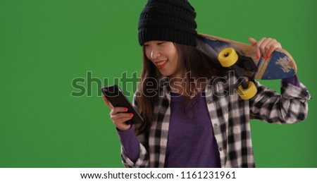 Asian skater girl carrying skateboard on shoulder using phone on green screen