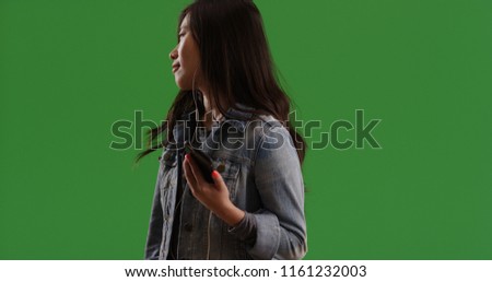 Hip Asian female in denim jacket listening to music on phone on green screen