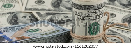 A roll of one hundred dollar bills tied with a rope against the backdrop of scattered dollars. Side view.
