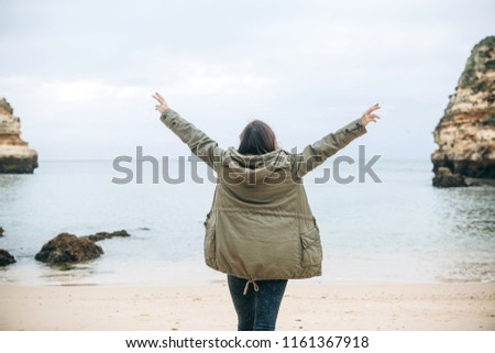 View from the back. A young woman tourist enjoys the beautiful views of the Atlantic Ocean and the landscape off the coast in Portugal and raises her arms upward showing how pleased she is.