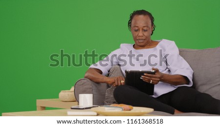 Elderly black woman uses her tablet while relaxing on the couch on green screen