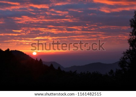 Alpine Mountain Sunset, deep red warm colors, rays of sunshine. Sun beginning to set below clouds in the distance, forest silhouette in the foreground. Taiwan Alishan mountain national forest