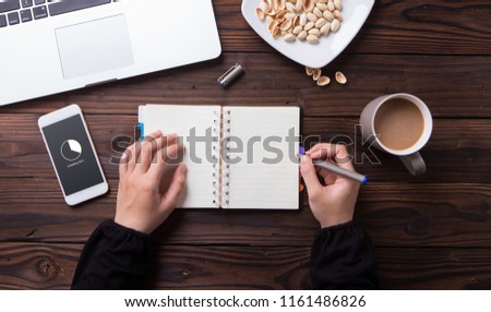 While waiting for installing on smartphone a woman write something on notebook with wooden desk background